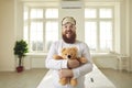 Happy grown-up man in pajamas standing in bedroom and cuddling teddy bear like a baby