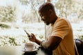 Happy cheerful African hipster holding mug, drinking fresh cappuccino, browsing internet and checking newsfeed on social