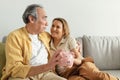 Happy cheerful european senior couple holding piggy bank, sitting on sofa at home, selective focus Royalty Free Stock Photo