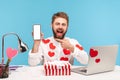 Happy cheerful man with beard in white shirt with red heart shaped stickers pointing finger at white display of smartphone in his Royalty Free Stock Photo