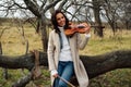 Charming woman in coat with violin sitting on a low tree branch at autumn