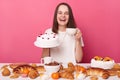 Happy cheerful woman wearing white T-shirt sitting at festive table with various desserts, isolated over pink background drinking Royalty Free Stock Photo