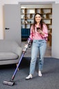 Happy cheerful young Caucasian beautiful woman vacuuming floor and texting, surfing internet on smartphone at home in living room Royalty Free Stock Photo