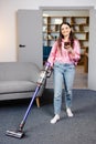 Happy cheerful young Caucasian beautiful woman vacuuming floor and texting, surfing internet on smartphone at home in living room Royalty Free Stock Photo