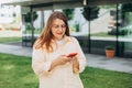 Happy cheerful young woman walking on city street checks her smartphone. Portrait of beautiful 30s businesswoman using Royalty Free Stock Photo