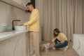 Happy cherrful couple in the kitchen in yellow hoodies, man washing the dishes and woman sitting and petting little white dog