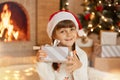 Happy child girl with Christmas present looking directly at camera, wearing red santa claus hat, holding gift box with ribbon in Royalty Free Stock Photo