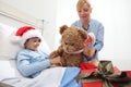 Happy child lying in bed in hospital room with Santa Claus hat and nurse dressing a teddy bear during Christmas holiday