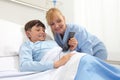 Happy child lying in bed in hospital room and smiling nurse using smartphone surfs the internet wearing earphones