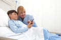 Happy child lying in bed in hospital room and smiling nurse using smartphone surfs the internet wearing earphones