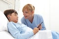 Happy child lying in bed in hospital room and smiling nurse using smartphone surfs the internet wearing earphones