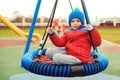 Happy child at outdoors playground. Funny kid boy having fun at park. Smiling boy swinging on modern swing. Child wearing red Royalty Free Stock Photo