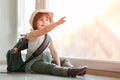 A happy child is ready for the journey. A baby with a backpack on her back is sitting at the window in the apartment. She put her Royalty Free Stock Photo