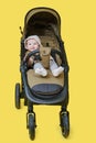 A happy child is sitting in a baby carriage on a studio yellow background. Smiling toddler baby boy in a transformer stroller for