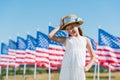 Child standing in white dress and touching straw hat near american flags