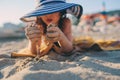 Happy child in swimsuit relaxing on the summer beach, lying on towel and playing with sand. Warm weather, cozy mood. Traveling on