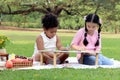 Happy children having a picnic in summer park, cute curly hair African girl with Asian buddy friend studying while sitting on mat Royalty Free Stock Photo