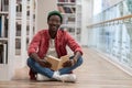 Happy clever black student man with book sitting on floor in university library, looking at camera Royalty Free Stock Photo