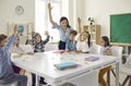 Happy school children and teacher sitting around classroom table and raising hands