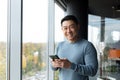 Happy and confident Asian businessman looking at camera and smiling, in modern office workplace, man holding phone Royalty Free Stock Photo