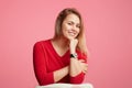 Happy confident blonde attractive woman keeps hand under chin, has shining smile, wears red sweater, isolated over pink background