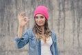 Happy confident smiling woman without make-up dressed in jeans shirt, white hat, with long curly hear showing ok-sign against grey Royalty Free Stock Photo