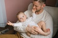 Happy contented father holds his baby son in his arms and sits at home in an armchair