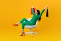 Happy contented shopaholic woman sitting on chair and raising hands with shopping bags, yellow background, full length