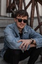 Happy cool young man hipster in stylish blue denim clothes in trendy sunglasses sits on a vintage staircase in the city