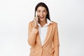 Happy corporate woman answer phone call, smiling and looking amazed, standing in suit over white background Royalty Free Stock Photo