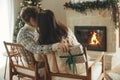 Happy couple in cozy sweaters kissing and exchanging stylish christmas gifts on background of fireplace with festive mantle and