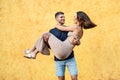 Happy couple on a date. Handsome man carrying his beautiful woman on hands. Yellow wall. A lot of copy space Royalty Free Stock Photo