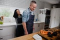 Happy couple laughing while wife ties on apron on husband standing in kitchen preparing dinner. Royalty Free Stock Photo