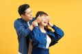 Happy Couple Playing Hide And Seek, afro man standing behind his wife, closing her eyes, yellow background