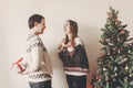 happy couple in stylish sweaters exchanging gifts in festive room at christmas tree. man holding surprise present behind back. me Royalty Free Stock Photo