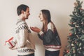 happy couple in stylish sweaters exchanging gifts in festive room at christmas tree. man holding surprise present behind back. me Royalty Free Stock Photo
