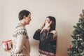 happy couple in stylish sweaters exchanging gifts in festive room at christmas tree. man holding surprise present behind back. me Royalty Free Stock Photo