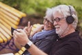 Happy couple of two  senior people white haired sitting on a yellow bench in video call with family with smart phone, bearded man Royalty Free Stock Photo