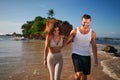 Happy couple walks barefoot on tropical beach at sunset, man leads woman by hand near ocean, romantic stroll by sea Royalty Free Stock Photo