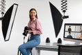 Happy creative photographer lady sitting on table workplace, holding her DSLR camera, working in modern photostudio