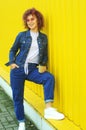 Happy curly haired woman in denim clothes drinking coffee on yellow background. Royalty Free Stock Photo