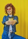 Happy curly haired woman in denim clothes drinking coffee on yellow background. Royalty Free Stock Photo