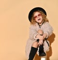 Little girl in sunglasses, pink fur coat, black hat and boots. Looking surprised, sitting cross-legged against beige background