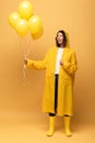 Happy curly woman in yellow raincoat and wellies holding balloons