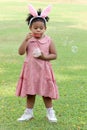 Happy cute African girl with black curly hair wearing bunny ears blowing soap bubbles in green nature garden. Kid spending time Royalty Free Stock Photo