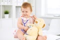 Happy cute baby at health exam at doctor`s office. Toddler girl is sitting and keeping stethoscope and teddy bear Royalty Free Stock Photo