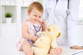 Happy cute baby at health exam at doctor`s office. Toddler girl is sitting and keeping stethoscope and teddy bear Royalty Free Stock Photo