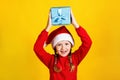 Happy cute baby holds a box over his head. Little girl in santa claus hat with christmas present on yellow background Royalty Free Stock Photo