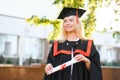 Happy cute caucasian grad girl is smiling. She is in a black mortar board, with red tassel, in gown, with nice brown Royalty Free Stock Photo