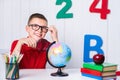 Happy cute clever boy is sitting at a desk in a glasses with raising hand. Child is ready to answer with a blackboard on Royalty Free Stock Photo